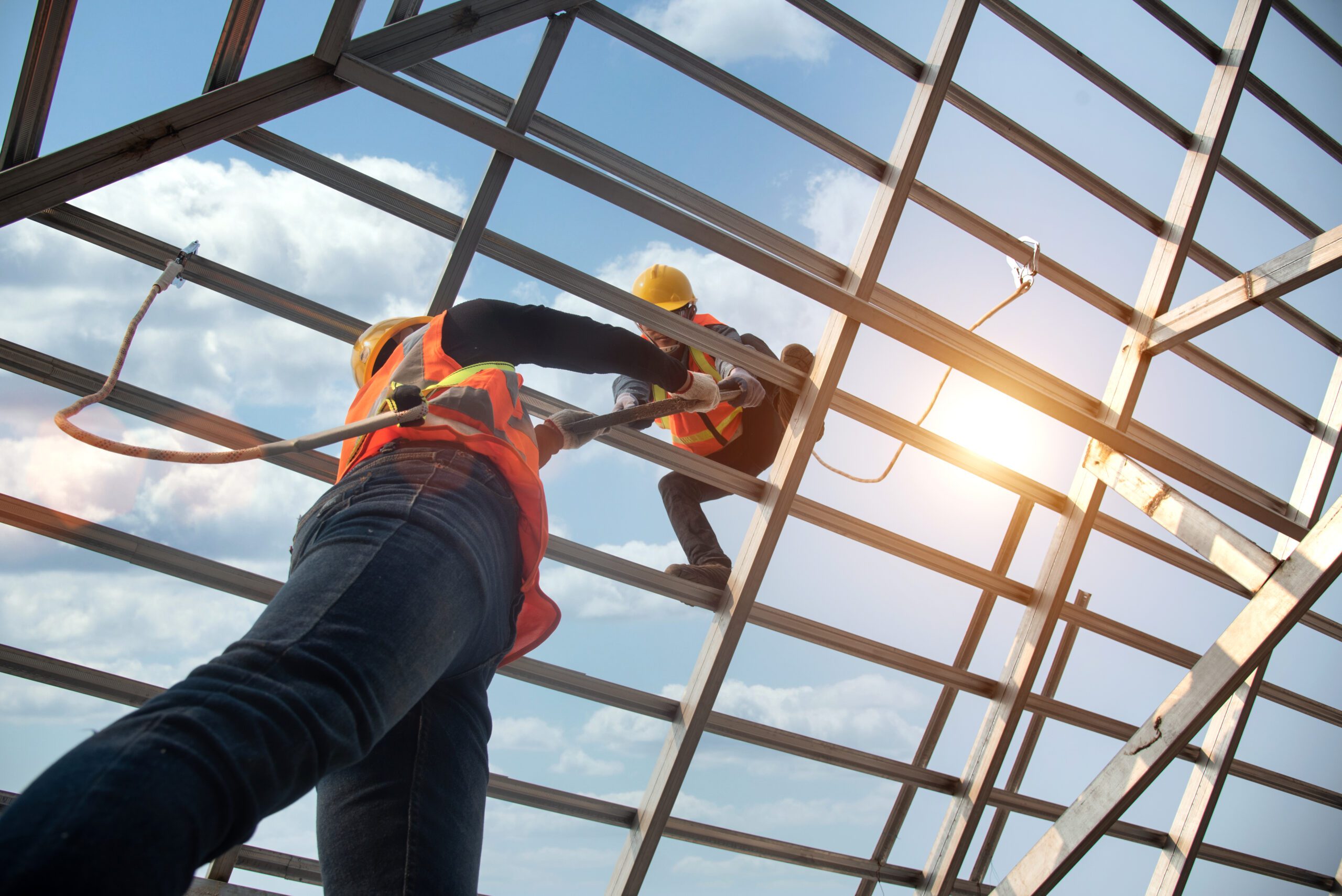 Two construction workers wearing safety gear are working on a metal structure with a blue sky and some clouds in the background.